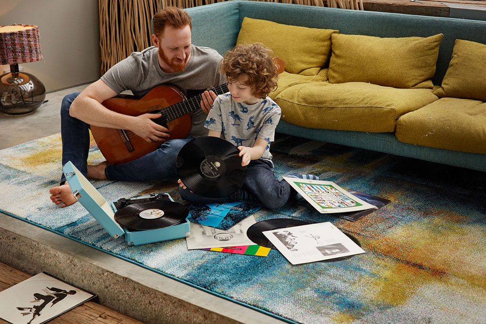 A photo of a father playing a guitar with his son playing with records